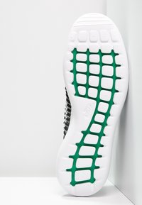 Biała podeszwa buta Nike z zielonym wzorem siatki, teksturowanym bieżnikiem oraz częścią czarno-biało górnej części widoczną na białym tle.