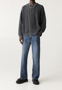 Pull gris texturé avec des motifs en losange, associé à un jean bleu à jambes droites et des chaussures noires. Accessoires comprenant un collier en chaîne argentée.