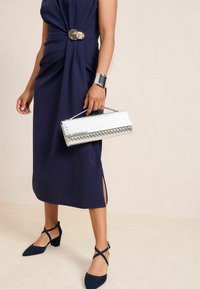 Meresinine kleit keeratud disaini ja kuldse aksendiga vöökohal. Sädelev hõbedane käekott geomeetrilise mustriga. Mustad plokksed kontsakingad.