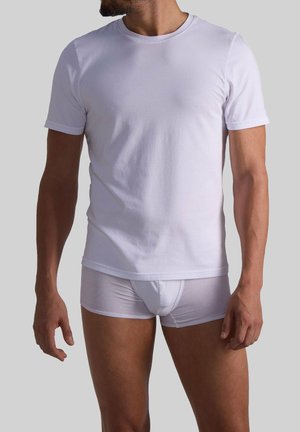 Weißes Baumwoll-T-Shirt mit kurzen Ärmeln und rundem Ausschnitt, kombiniert mit durchsichtigen weißen Unterhosen, getragen von einer Person im Profil.