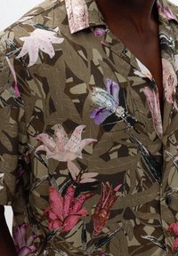 Kurzarmhemd mit khakifarbigen Hintergrund, das rosa, rote und lila Blumen- und Insektenmuster zeigt, getragen von einer Person.