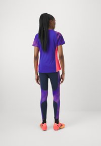 Conjunto atlético com uma parte superior de mangas curtas roxa e rosa, combinada com leggings azul marinho que apresentam um padrão texturizado roxo. Sapatilhas laranja brilhante.