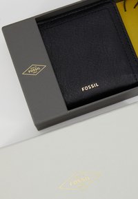 Zwart lederen portemonnee in een grijze doos, met een gouden reliëflogo. De buitenkant van de doos heeft een gestructureerde afwerking met een gele binnenkant.