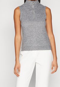 Mouwloos grijs gebreid topje met een hoge kraag en een zilveren rits bij de halslijn, voorzien van een geribde zoom en een gestructureerd patroon.
