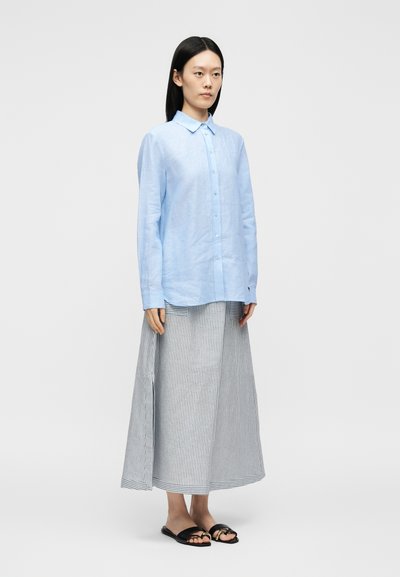 Femme debout portant une chemise bleu clair boutonnée, une longue jupe rayée grise et blanche, et des sandales noires à bouts ouverts sur un fond uni.