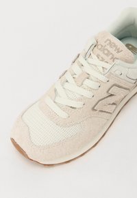 Baskets New Balance beige et blanches en daim et mesh, lacets blancs et semelle en gomme, inclinées pour montrer les détails du dessus et du côté.