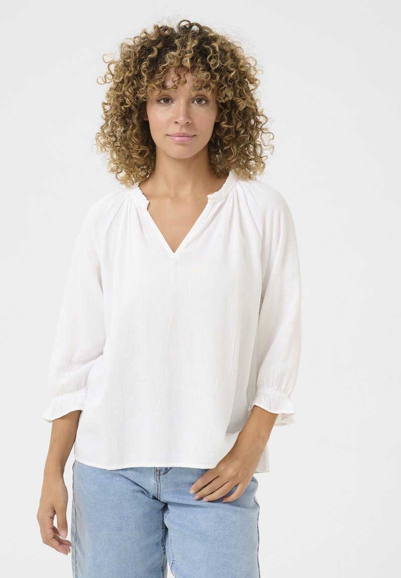 Blusa bianca leggera con scollatura a V, maniche a sbuffo con polsini arricciati e vestibilità comoda, abbinata a jeans in denim blu. Tessuto liscio.