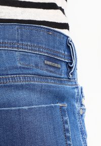 Blå denimjeans med en strukturerad yta, har en synlig logotyp-tag, fem fickor och kontrasterande sömmar längs sömmarna.
