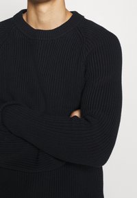 Pull en maille côtelée bleu foncé avec col rond et manches longues, présentant un motif texturé et une coupe décontractée.