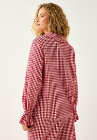 Woman with curly hair wears a red and white checkered long-sleeve shirt and matching skirt, standing turned slightly to the side.