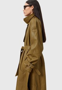 Olijfgroene leren trenchcoat met een riem, grote kraag en zijzakken. Heeft een gladde textuur en manchetten met metalen accenten.