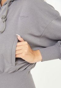 Felpa grigia chiara con finitura testurizzata, dotata di cappuccio con lacci, polsini a coste e tasca laterale con zip. Logo argento sul petto.