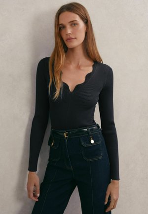 Haut noir à manches longues côtelé avec un col festonné. Associé à un jean en denim foncé taille haute et une ceinture noire avec des accents dorés.