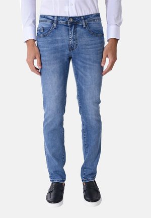 Uomo che indossa jeans slim fit blu, camicia bianca e scarpe slip-on nere con suola bianca, in piedi davanti a uno sfondo semplice.