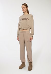 Felpa accorciata beige e pantaloni della tuta abbinati con il logo "KENTUCKY" in marrone scuro. Materiale morbido, vestibilità rilassata, abbinati a stivali marroni.