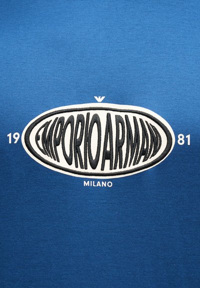 Μπλε ύφασμα με λευκό οβάλ μπαλώμα που φέρει εκτεταμένο μαύρο κείμενο "EMPORIO ARMANI," αριθμούς "19" και "81" στα πλάγια, και "MILANO" από κάτω.