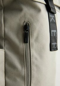 Sac en tissu beige avec une fermeture éclair lisse et des accessoires en métal noir, une surface texturée et une sangle noire avec des accents de marque blancs.