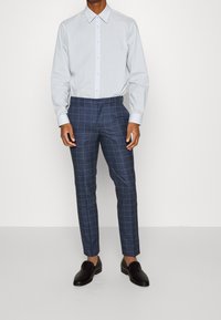 Hellblaues Langarmshirt mit Knopfleiste, kombiniert mit marineblauen karierten Hosen und schwarzen strukturierten Loafers, vor einem schlichten Hintergrund stehend.