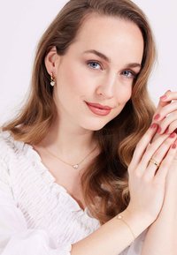 Goldschmuck umfasst eine herzförmige Anhängerkette, passende Ohrringe und ein Armband, das mit klaren Edelsteinakzenten verziert ist, vor einem strukturierten, hellen Hintergrund.
