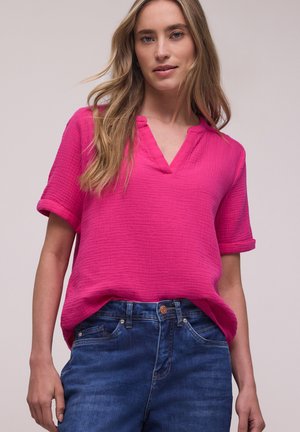 Mujer con cabello largo y rubio, con una blusa de manga corta texturizada de color rosa brillante y jeans de mezclilla azul, sobre un fondo liso.