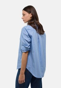 Blouse rayée bleu clair avec manches retroussées, coupe décontractée, patte de boutons dans le dos et ourlet arrondi. Associée à un jean foncé.