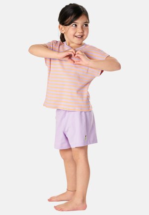 Young girl in striped orange and purple shirt and purple shorts making a heart shape with her hands, smiling and looking to the side.