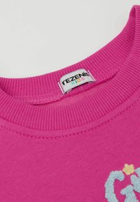 Růžová mikina vyrobená z měkké bavlny, s vroubkovaným kulatým výstřihem a vyšitým vzorem v modré a žluté barvě na přední straně.