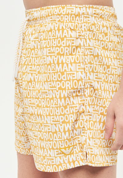 Pantaloni da bagno gialli con una ripetizione di un motivo testuale bianco che presenta "Emporio Armani". Vita elastica con coulisse, dettaglio del logo sull'orlo.