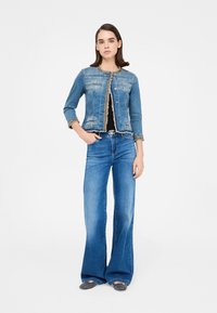 Jeans jakna svetlo modre barve z okrasnim zlatim trakom ob ovratniku in manšetah, kombinirana z modrimi kavbojkami širokih hlačnic.