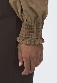 Top marrón de manga larga con puño fruncido y borde con volantes, combinado con una prenda ajustada marrón oscuro. Detalle de tela texturizada visible.