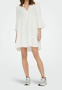 Witte jurk tot op de knie met een V-hals, uitlopende mouwen en een ontspannen pasvorm; gedragen met grijze en witte sneakers voor een casual look.