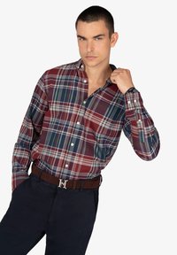 Camisa de cuadros roja, azul y verde, de botones, de manga larga, confeccionada en algodón, llevada con un cinturón marrón que presenta una hebilla de plata grabada.