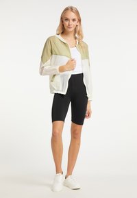 Letvægts, farveopdelt jakke i olivengrøn og hvid, med lynlås og elastiske manchetter, parret med sorte cykelshorts og hvide sneakers.