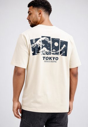 Man wearing beige T-shirt with black graphic panels of waves, Mount Fuji, and a sun, and text "TOKYO" on the back.