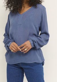 Frau mit lockigem Haar, die eine lockere blaue Langarmbluse und blaue Jeans trägt, mit goldenen Ringen an ihren Fingern vor einem neutralen Hintergrund.