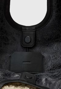 Zwarte leren tas met een textuur, voorzien van een ronde opening, een ronde knoopsluiting en een plat leren label met merknaam aan de onderkant.