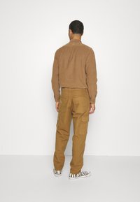 Rudas kadro kompleksas marškiniai, trumpesnio stiliaus, derinami su smėlio spalvos kargo kelnėmis, turinčiomis kelis kišenes. Avalynė: juodi ir balti raštuoti sportbačiai.