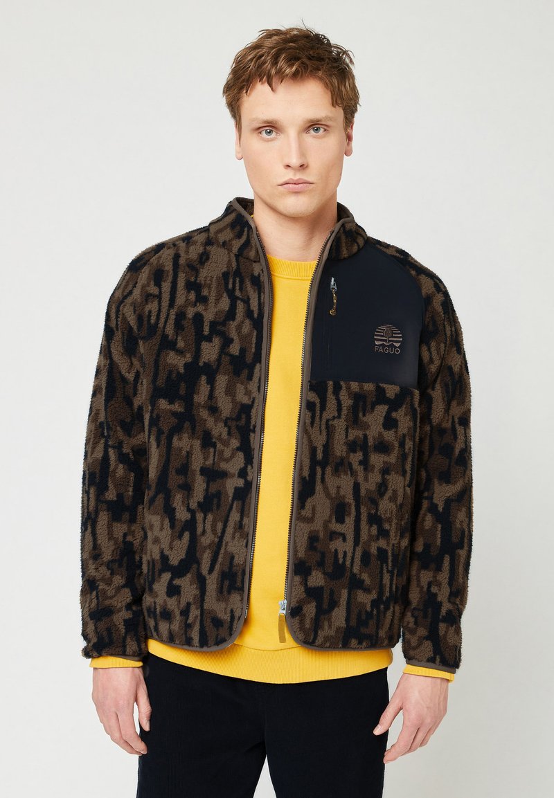 Giacca in pile marrone fuzzy con motivo astratto nero, zip completa e tasca blu navy. Indossata sopra una maglietta gialla, presenta un logo sul petto.
