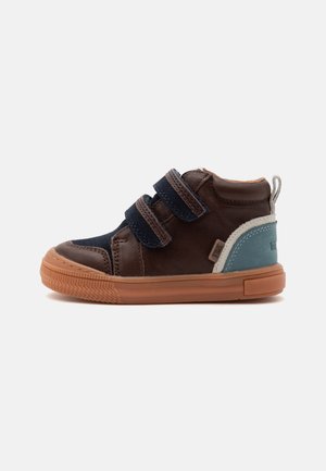Chaussure pour tout-petit marron avec brides en velcro bleu marine, semelle en caoutchouc beige et patch bleu et blanc au talon sur fond blanc.