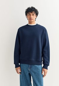 Felpa blu navy in misto cotone, con scollo rotondo, polsini e orlo a coste, dalla vestibilità comoda. Indossata con jeans azzurri.
