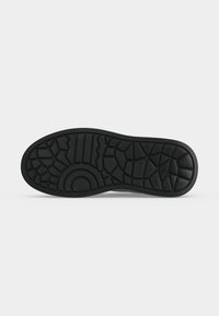 Semelle de chaussure noire avec un motif géométrique de la bande de roulement comprenant des arcs, des zigzags et des formes polygonales, présentée sur fond blanc.