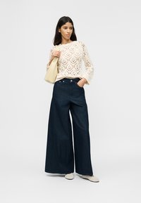 Γυναίκα που φοράει κρεμ πλεκτό πουλόβερ, φαρδιά σκουρόχρωμα τζιν, λευκά παπούτσια και κρατάει μια μπεζ τσάντα στον ώμο, στέκεται μπροστά σε απλό φόντο.