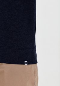Navy blauwe sweatshirt met geribbelde onderkant en manchetten, gemaakt van structuurstof. Bevat een klein wit logo-label aan de onderzijde links.