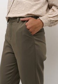 Pantalon vert olive avec poches latérales, coupe ajustée, tissu lisse, doté d'une ceinture et d'un léger éclat. Associé à un haut clair texturé.