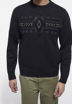 Mann trägt schwarzen Pullover mit strukturiertem geometrischem Muster über der Brust und olivgrüner Hose, gezeigt vom Hals bis zu den Hüften.