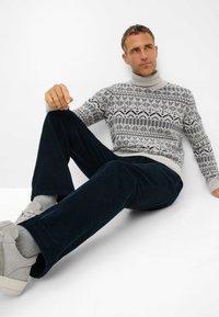 Maglione grigio chiaro con motivo e colletto alto, abbinato a pantaloni in velluto a coste blu scuro e sneakers grigio chiaro. Texture liscia in tutto il capo.