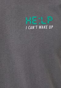 Grå bomulds t-shirt med grøn tekst i digital stil: "HE:LP" over hvid tekst, der siger "JEG KAN IKKE VÅGNE OP." Enkel, afslappet design.