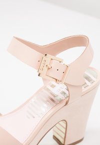 Sandale à talons aiguilles rose avec une finition brillante, dotée d'une boucle dorée, d'une bride de cheville et d'une semelle intérieure texturée avec un motif rayé.