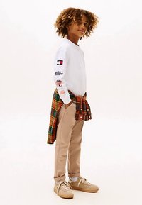 Hvit langermet skjorte med grafiske detaljer, beige bukser, beige joggesko og en rutete skjorte i grønt, rødt og gult knytt rundt livet.