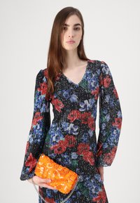 Robe noire à motifs floraux avec de longues manches bouffantes et un décolleté en V. Tenant une pochette en sequins orange avec une bandoulière en chaîne argentée.